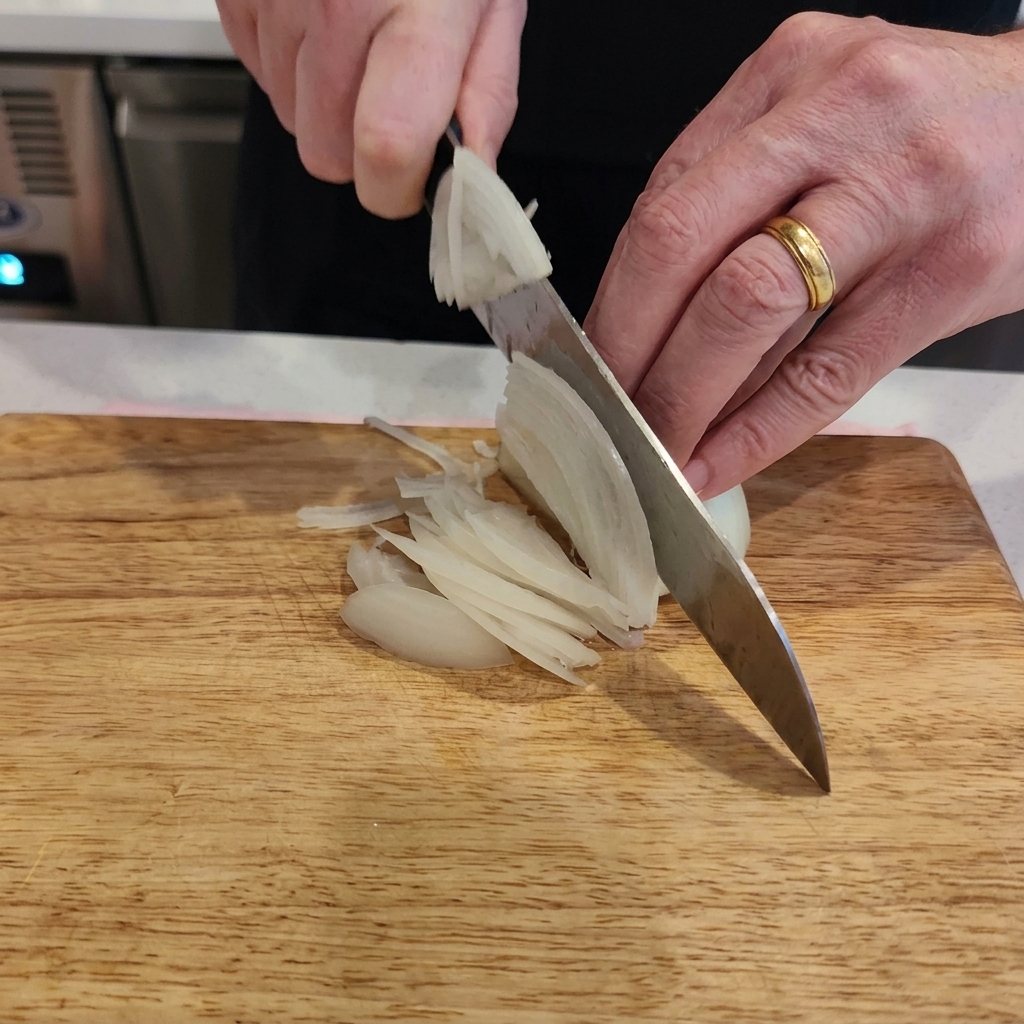Gros plan sur des mains coupant un oignon blanc en fines lamelles avec un couteau de chef sur une planche en bois.