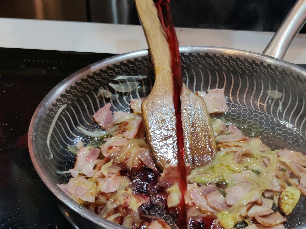 Un filet de vin rouge foncé versé depuis un verre dans une poêle en acier inoxydable contenant les lardons et les oignons crépitants.