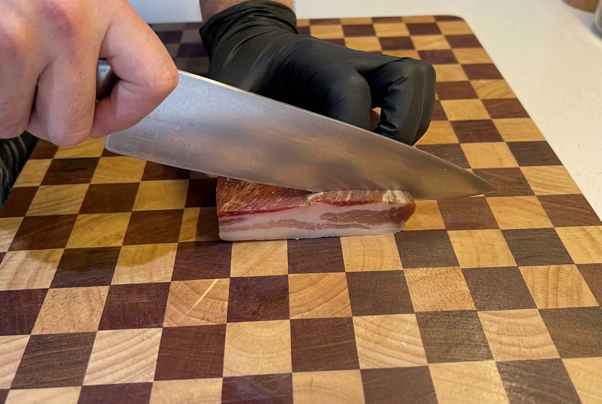 Mains avec des gants noirs utilisant un grand couteau de chef pour couper un morceau épais de pancetta en petits dés sur une planche en bois.