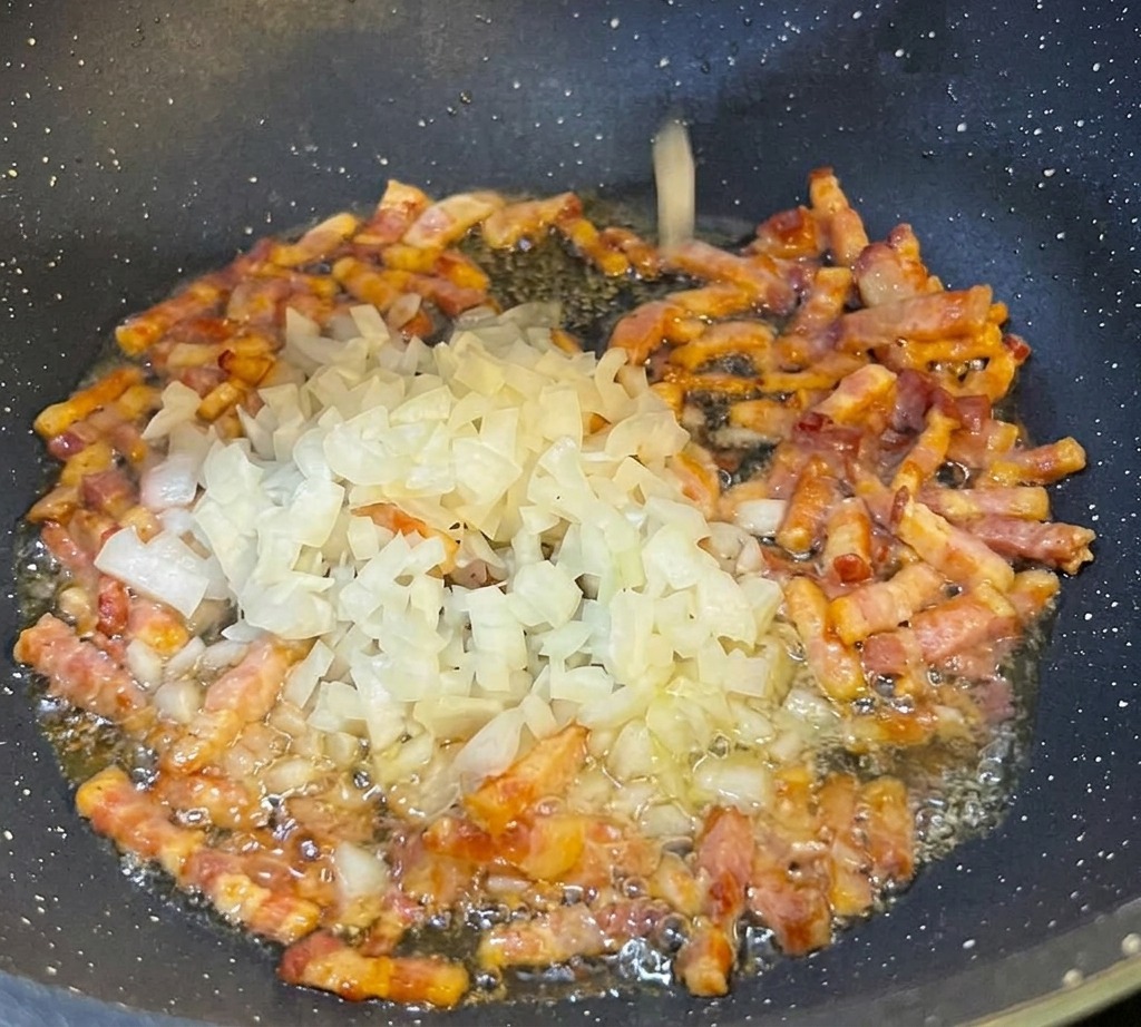 Oignons blancs finement émincés parsemés dans une poêle noire contenant des lardons de pancetta en train de dorer.