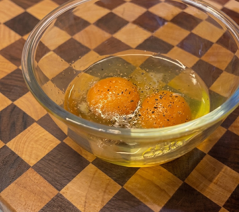 Une personne moulant du poivre noir avec un moulin en bois dans un bol en verre contenant deux jaunes dœufs orange vif.