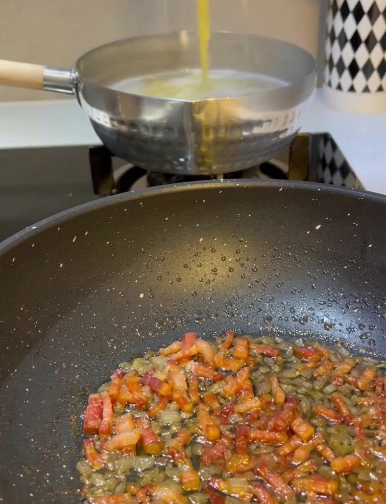 Une main utilisant des pinces métalliques pour attraper les spaghetti jaunes dune casserole deau bouillante et les transférer dans une poêle contenant la pancetta et les oignons cuits.