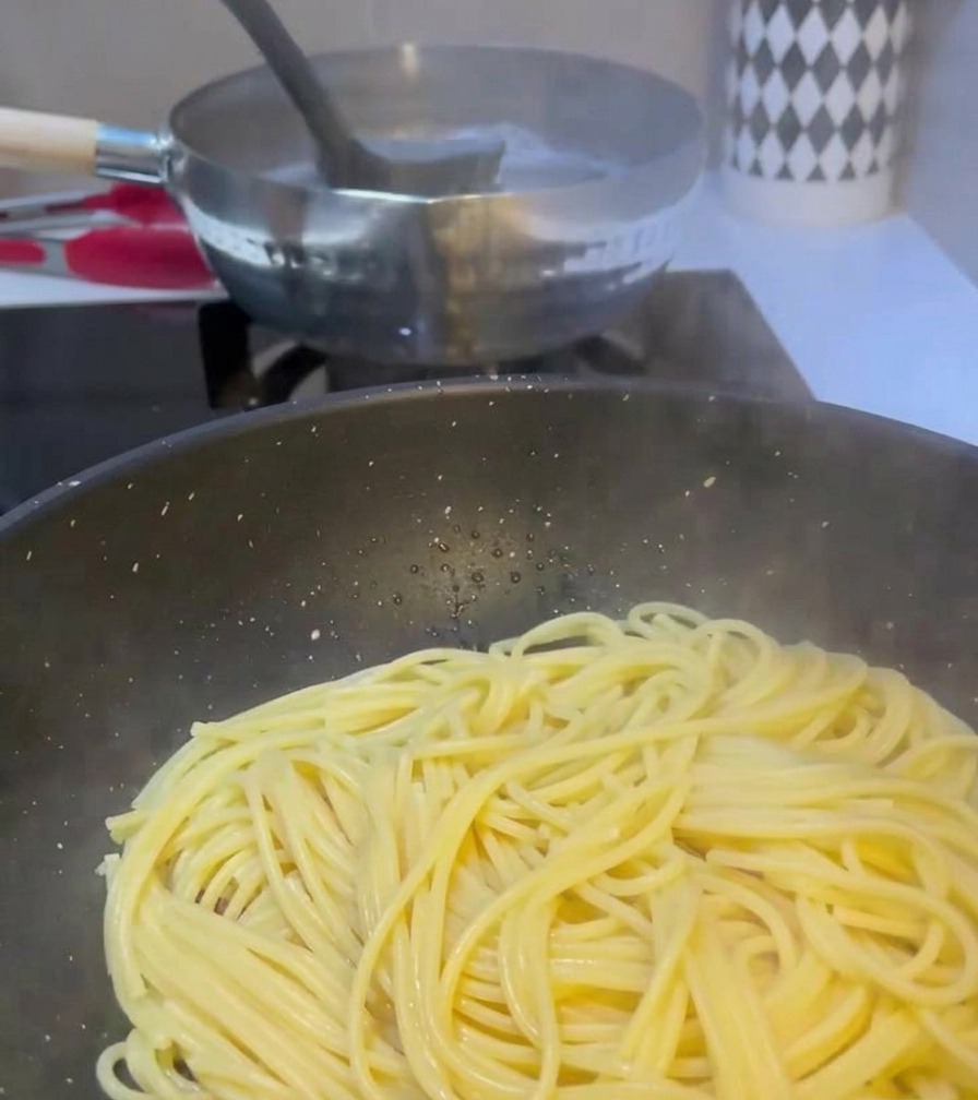 Une louche transférant de leau trouble dune casserole de pâtes en ébullition vers une poêle contenant déjà des spaghetti et de la viande cuite.