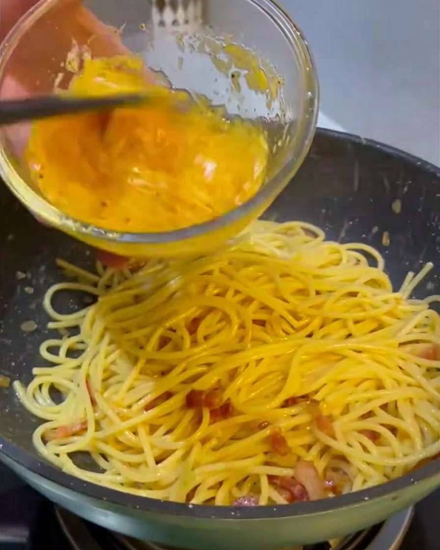 Verser un mélange jaune vif dœufs et de fromage depuis un bol en verre sur une poêle de spaghetti chauds et de viande frite.
