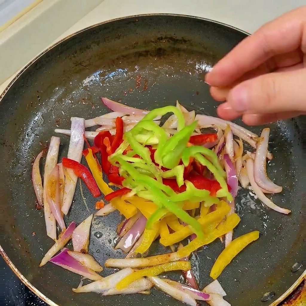Une main ajoutant des lanières de poivrons rouges, jaunes et verts dans une poêle contenant déjà des oignons revenus.