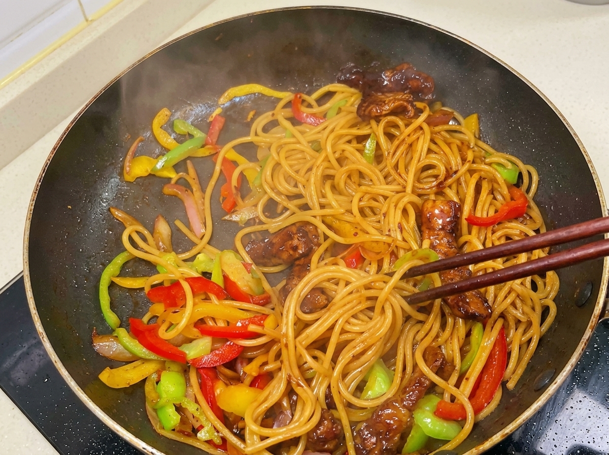 Des baguettes mélangeant activement des spaghettis fumants, du porc doré et des poivrons enrobés dune sauce sombre et brillante.