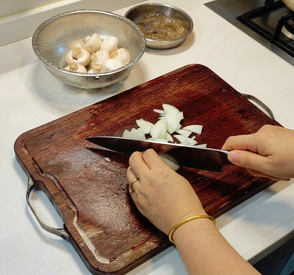 Mains coupant finement un oignon blanc sur une planche en bois sombre à côté de champignons.