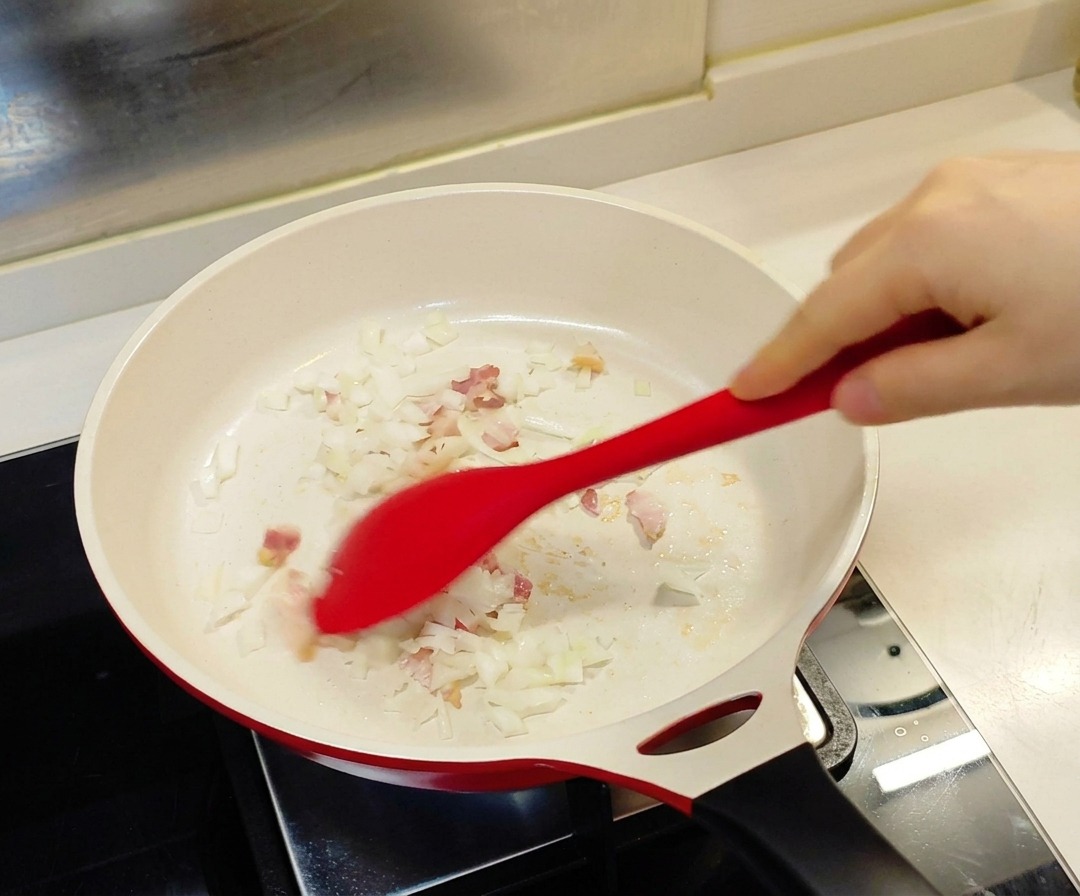 Une spatule rouge remuant des oignons émincés et des morceaux de bacon dans une poêle blanche.