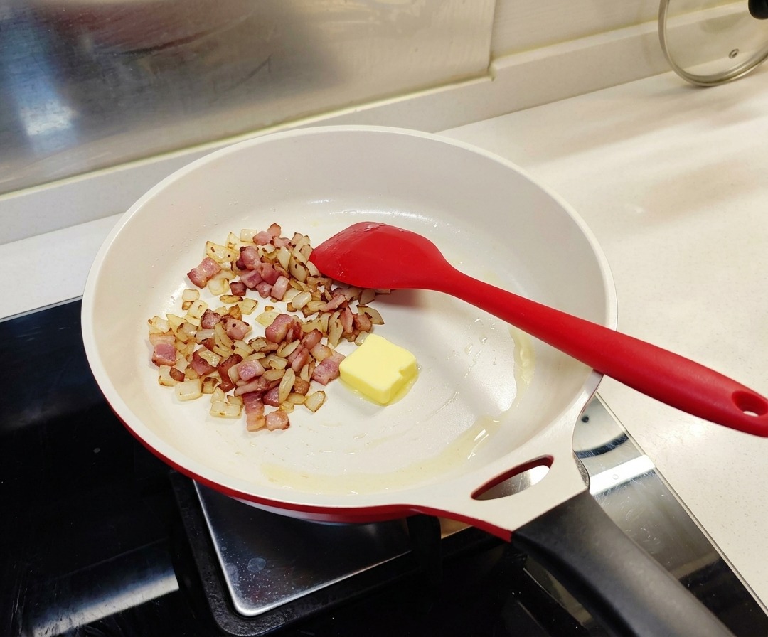 Un carré de beurre jaune fondant dans une poêle au milieu du bacon et des oignons cuits.