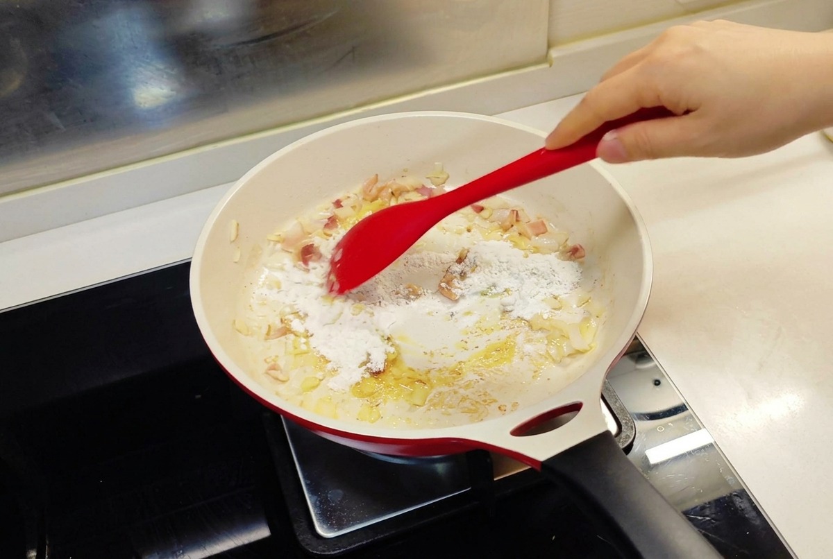 Une spatule rouge incorporant de la farine dans une poêle contenant des oignons et du beurre fondu.