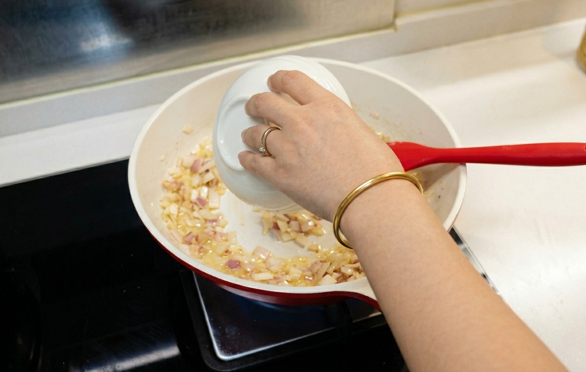 Une personne versant un peu deau depuis un bol blanc dans la poêle contenant le roux aux oignons.