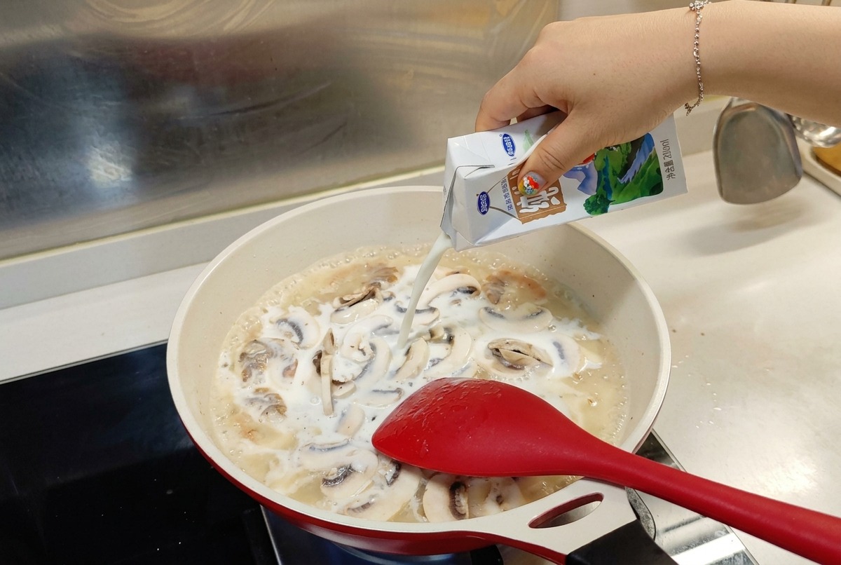 Du lait versé depuis une brique blanche et bleue dans la poêle où mijotent les crevettes et les champignons.