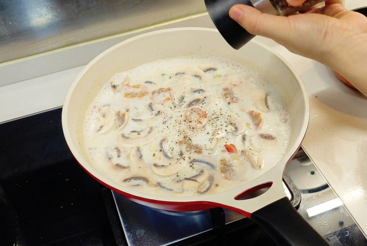 Une main utilisant un moulin pour ajouter du poivre noir dans la sauce blanche qui mijote dans une poêle rouge.