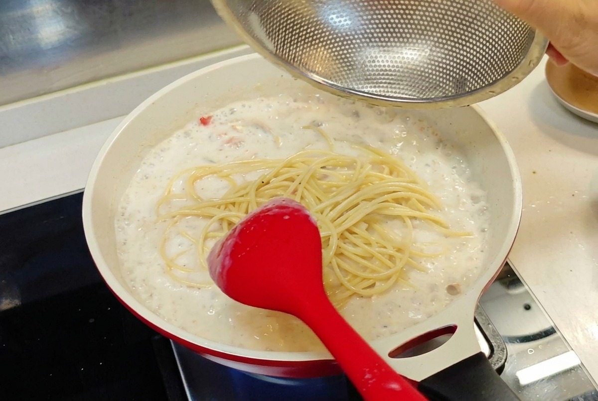 Spaghettis cuits transférés depuis une passoire fine dans la poêle de sauce blanche crémeuse.