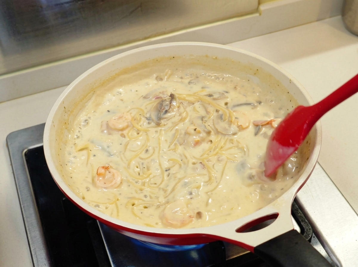 Une spatule rouge mélangeant des spaghettis en sauce crème avec crevettes et champignons dans une poêle en céramique.