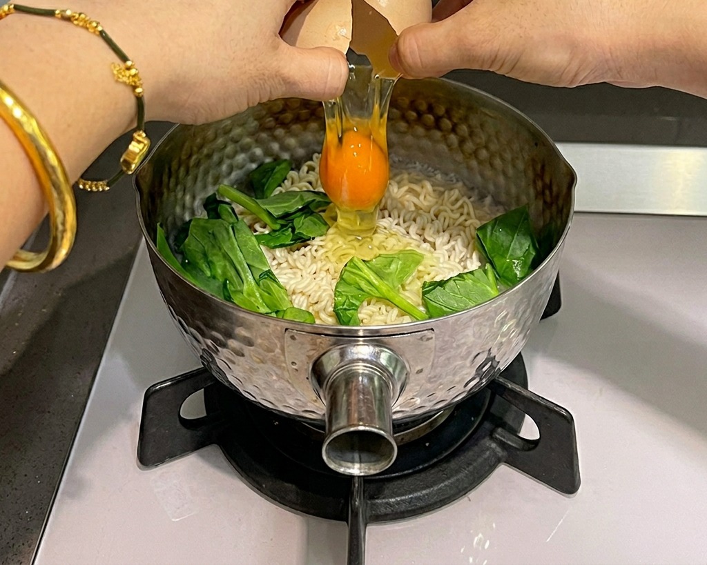 Deux mains cassent un œuf cru au-dessus dune casserole bouillante contenant des nouilles instantanées et des épinards.