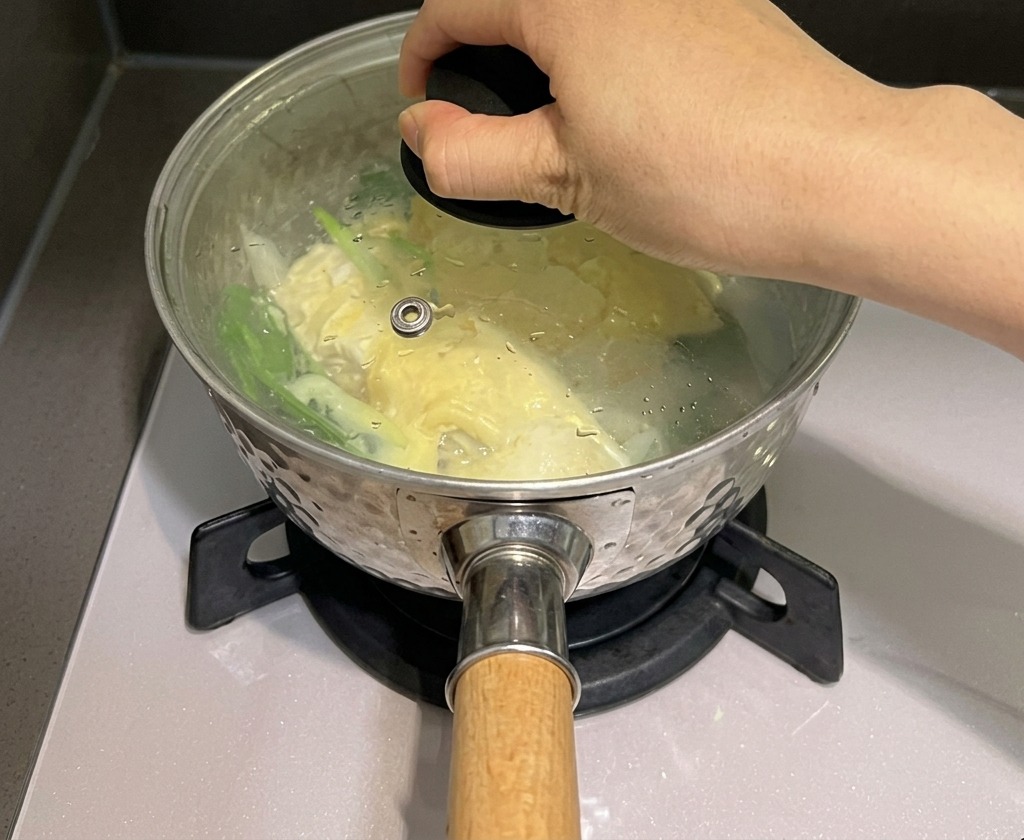 Une main place un couvercle en verre sur une casserole contenant des nouilles et du fromage pour emprisonner la vapeur.