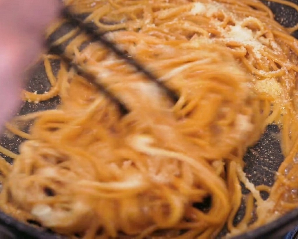 Plan daction de pâtes au blé complet mélangées vigoureusement avec des baguettes noires dans une poêle, montrant le fromage en poudre et le beurre fondu former une sauce.