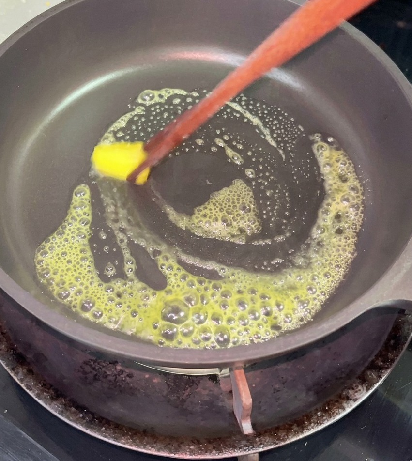 Un petit morceau de beurre jaune qui fond et mousse dans une poêle antiadhésive noire, remué avec des baguettes en bois.