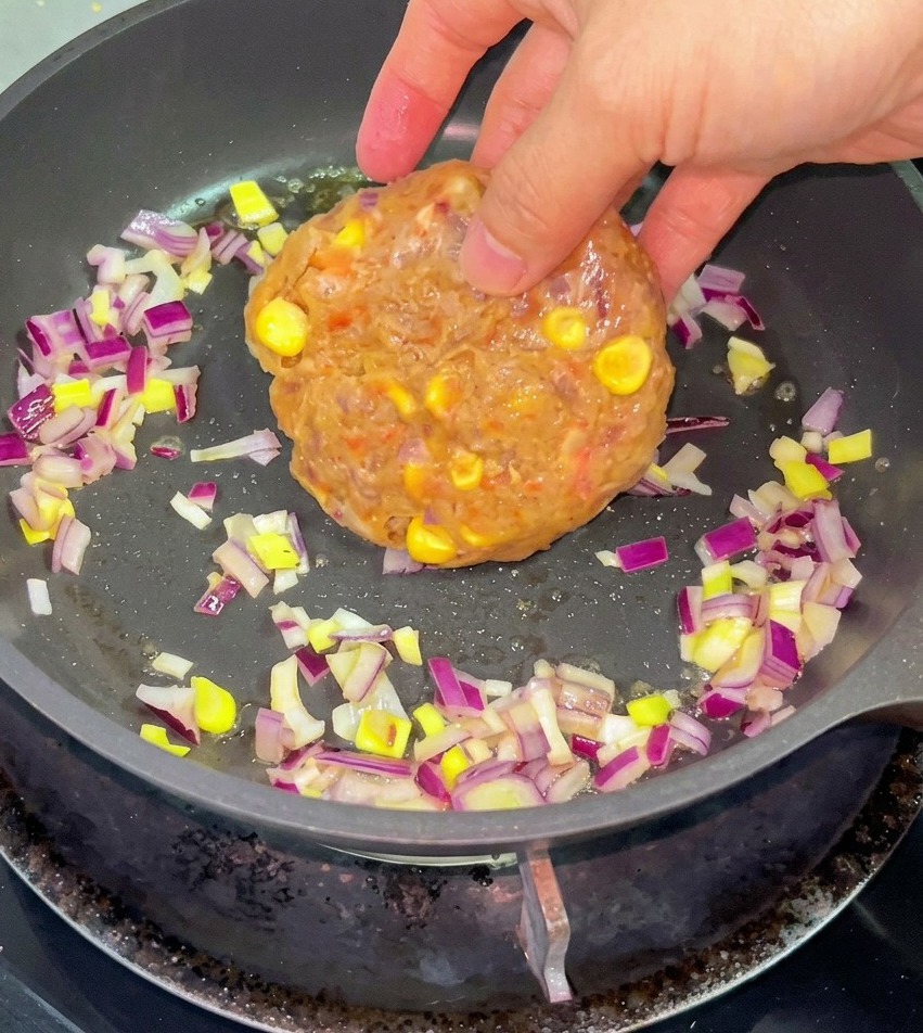Une main déposant une préparation de bœuf haché et de maïs dans une poêle avec des oignons rouges émincés et du beurre fondu.