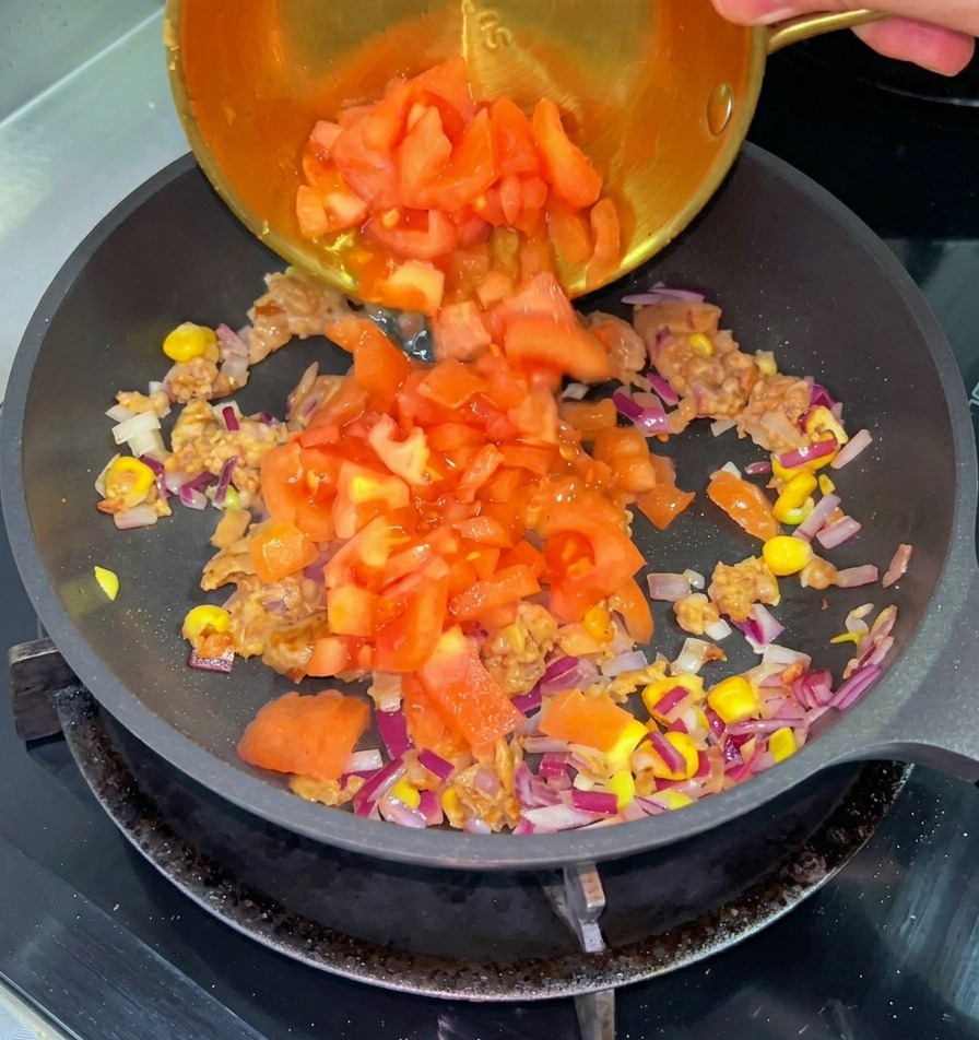 Des tomates rouges coupées en dés versées depuis un petit bol en laiton dans une poêle contenant le bœuf cuit, le maïs et les oignons.
