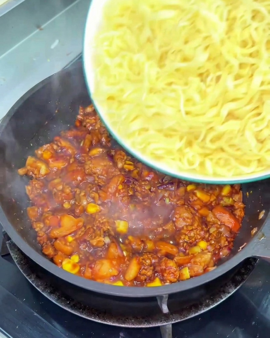 Un bol de nouilles jaunes ondulées versé dans une poêle remplie dune sauce épaisse à la tomate et au bœuf haché.