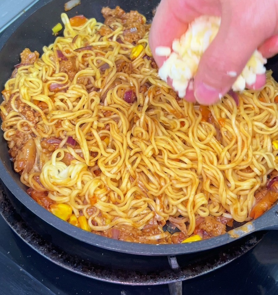 Gros plan dune main saupoudrant du fromage blanc râpé sur une poêle de nouilles sautées mélangées à du bœuf haché, des oignons et du maïs.