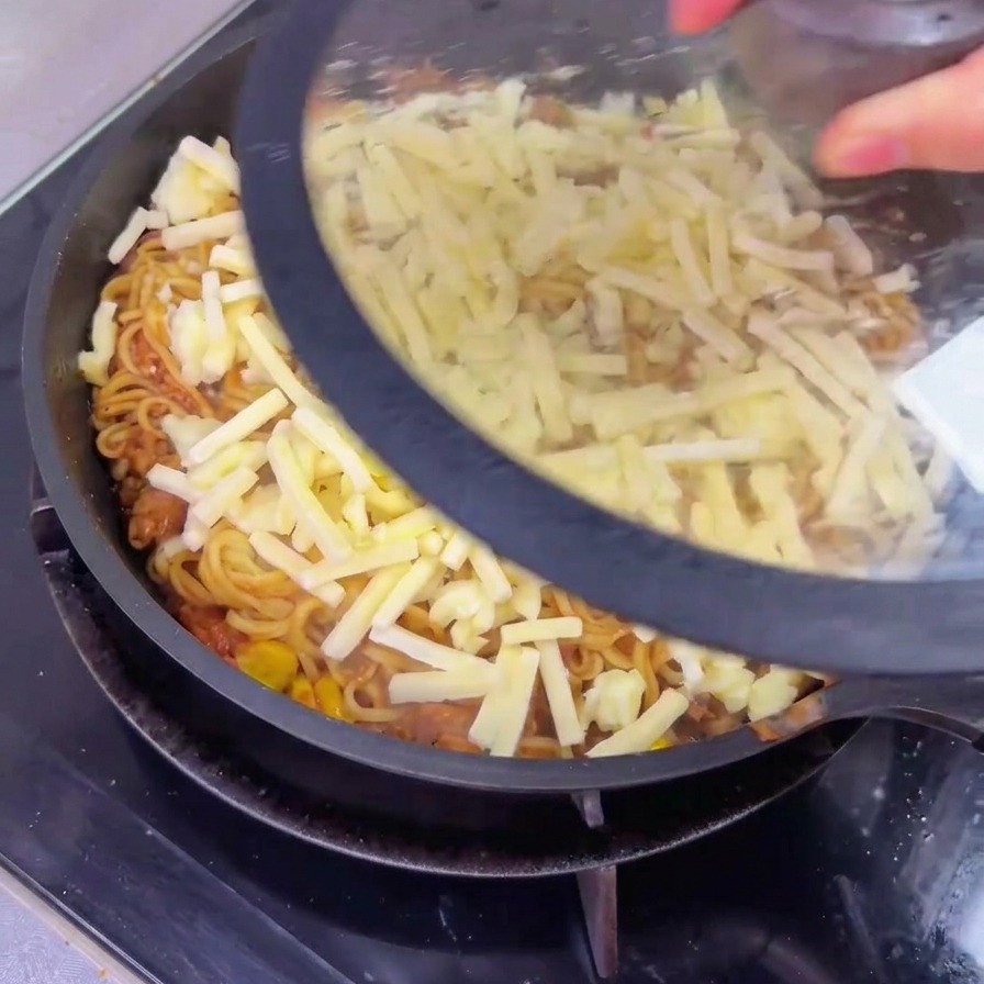 Une personne posant un couvercle en verre transparent sur une grande poêle contenant des nouilles au bœuf recouvertes de fromage râpé.