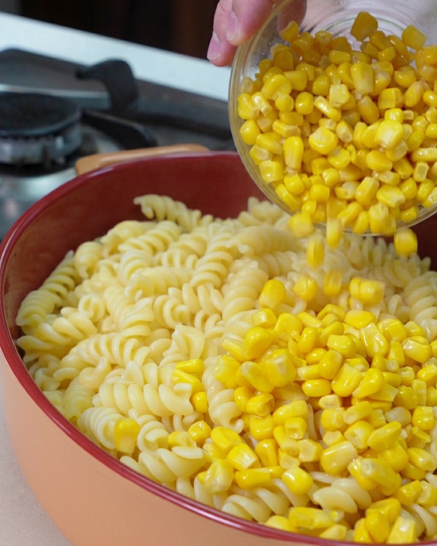 Un bol en verre versant des grains de maïs doux jaune vif sur un lit de fusillis cuits dans un plat à gratin rond.