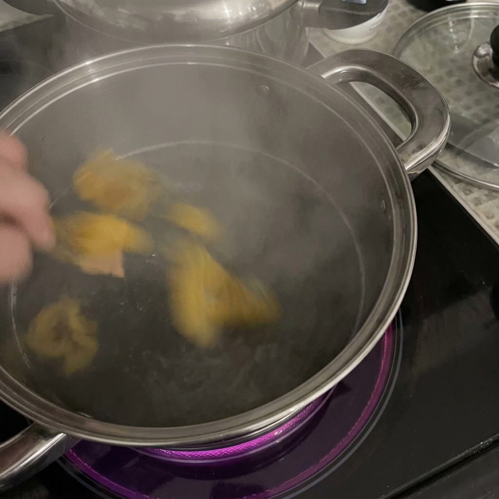 Une main plongeant des rigatoni jaunes dans une casserole en acier inoxydable remplie deau bouillante sur une plaque à induction.