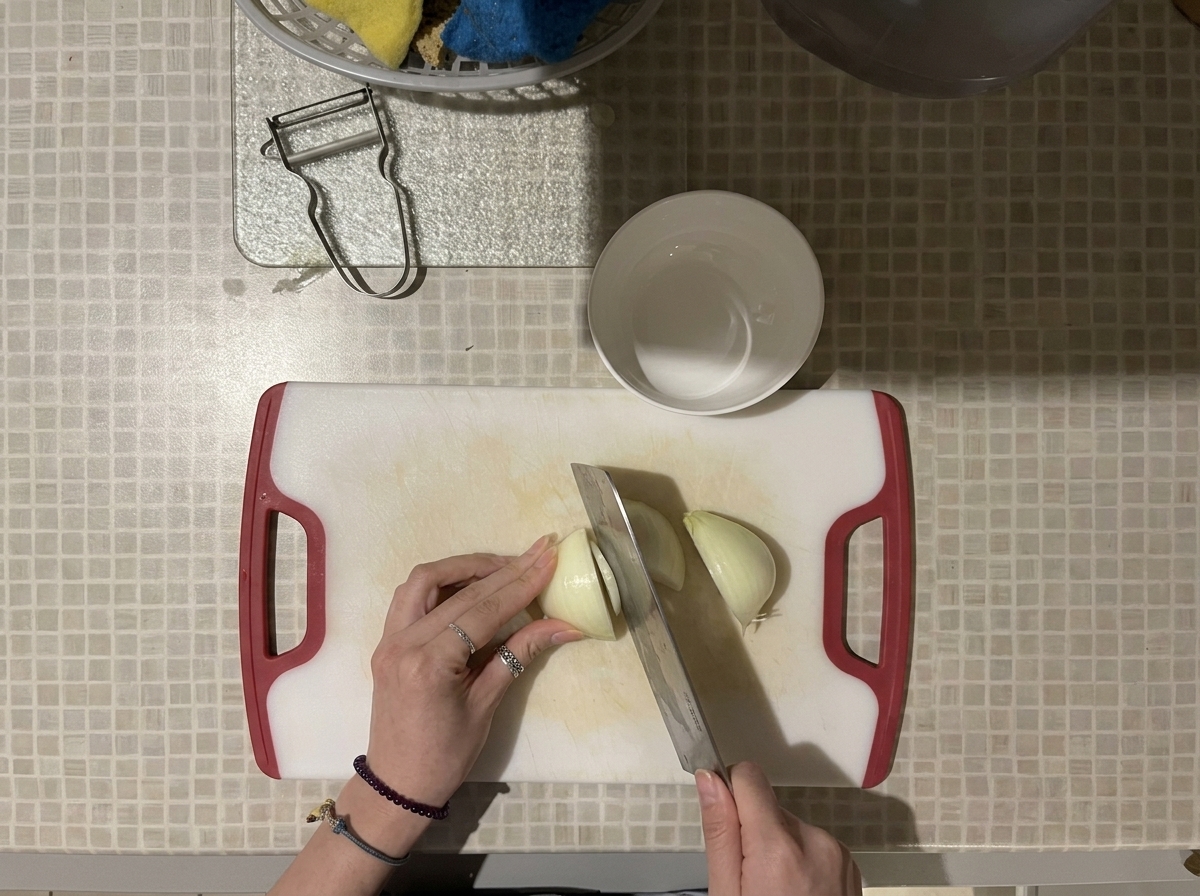 Vue du dessus de mains utilisant un grand couteau pour couper un oignon blanc en dés sur une planche à découper blanche aux poignées rouges.