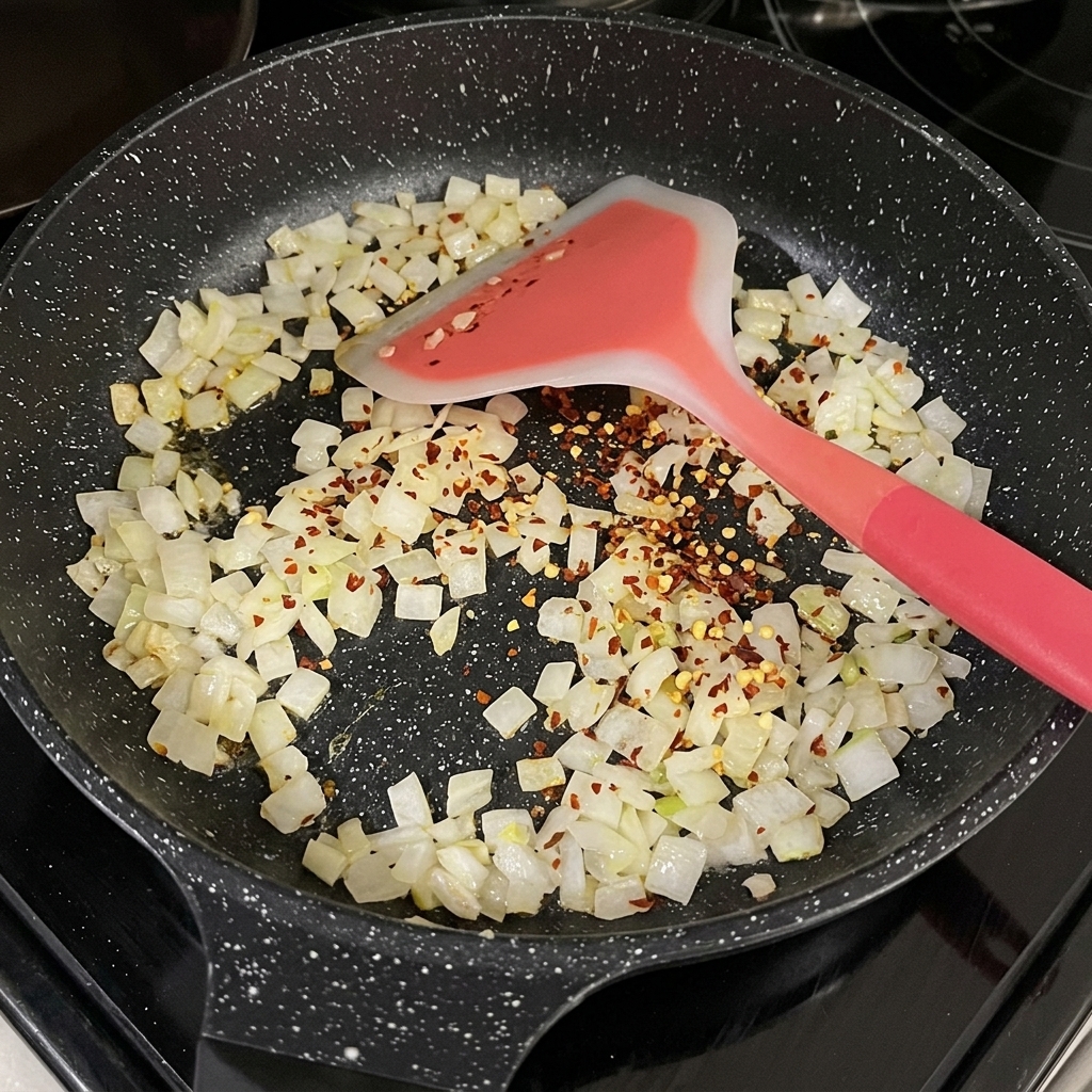 Flocons de piment rouge séché parsemés sur des oignons translucides en dés dans une poêle noire.