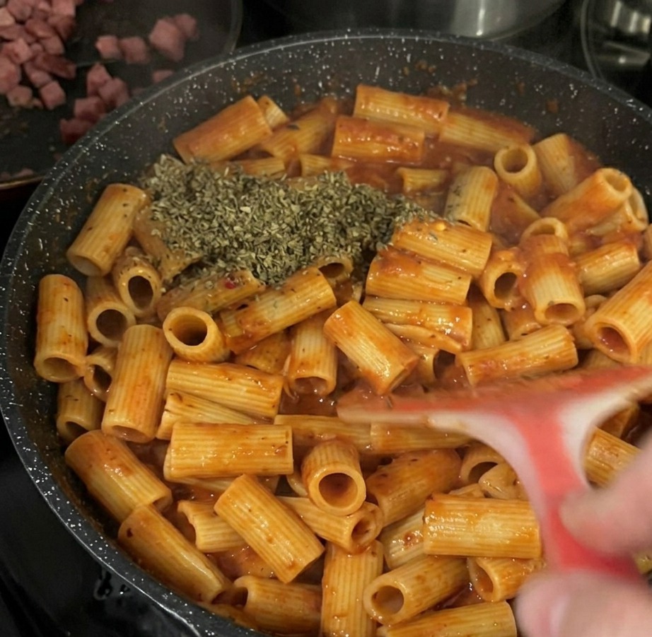 Une généreuse pincée de flocons de basilic séché vert ajoutée sur les rigatoni en sauce tomate dans une poêle noire.