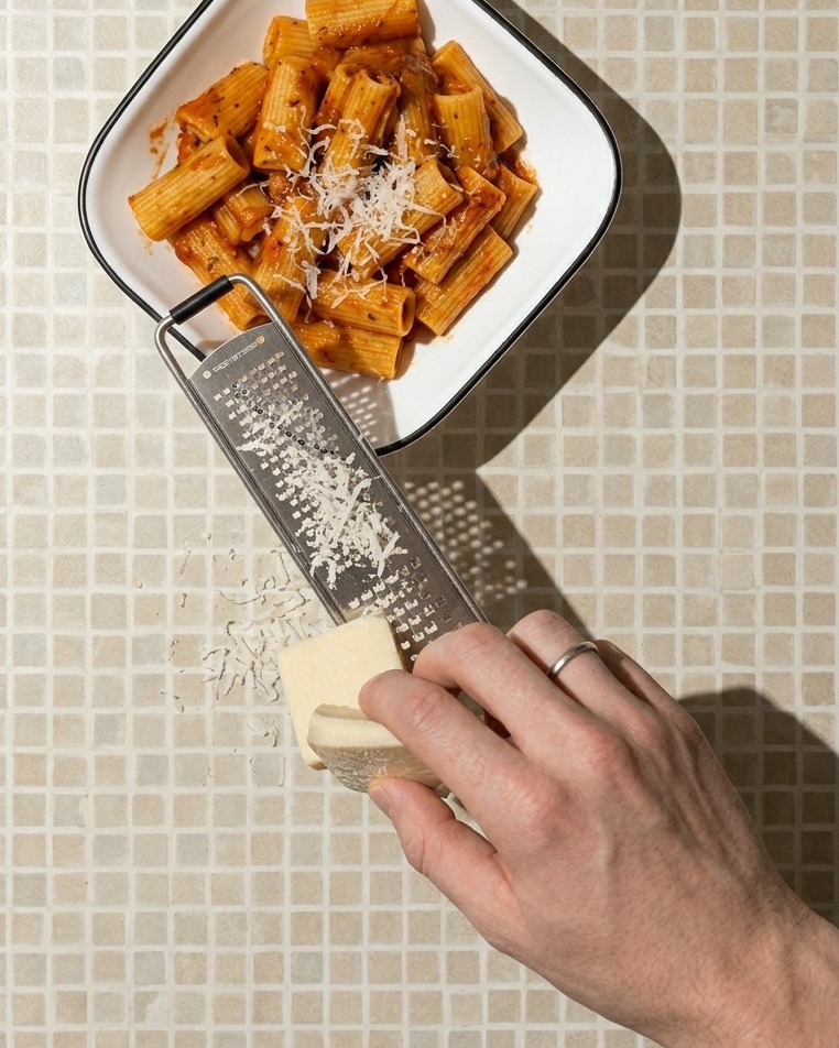 Gros plan dune main râpant un bloc de fromage blanc au-dessus dun bol carré blanc rempli de rigatoni à la sauce rouge.