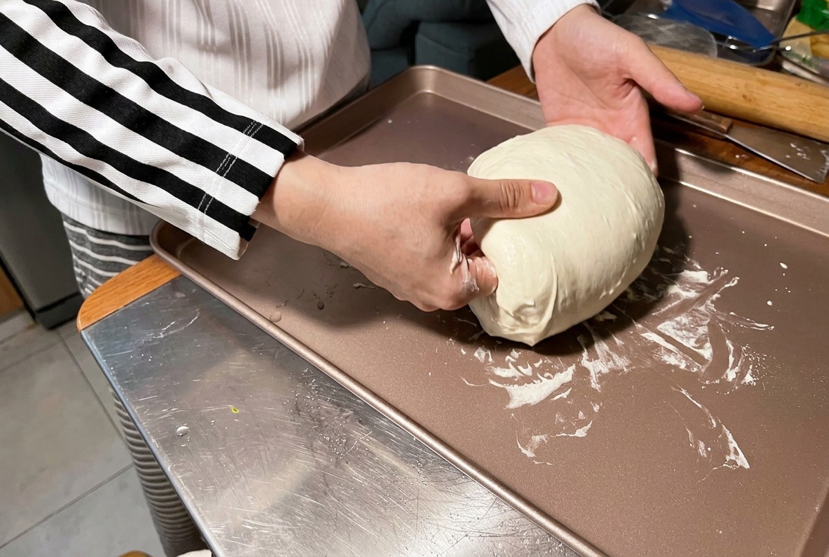 Une personne utilise ses mains pour bouler et lisser une grosse boule de pâte blanche et souple sur une plaque de cuisson de couleur bronze.