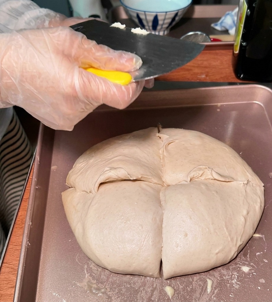 Une personne portant un gant en plastique utilise un coupe-pâte métallique pour dutiliser diviser une grande pièce de pâte en portions plus petites et arrondies sur un plateau.