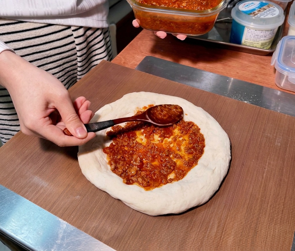 Une cuillère en bois étale une sauce tomate épaisse et rouge à la viande en effectuant un mouvement circulaire sur un disque de pâte à pizza crue.