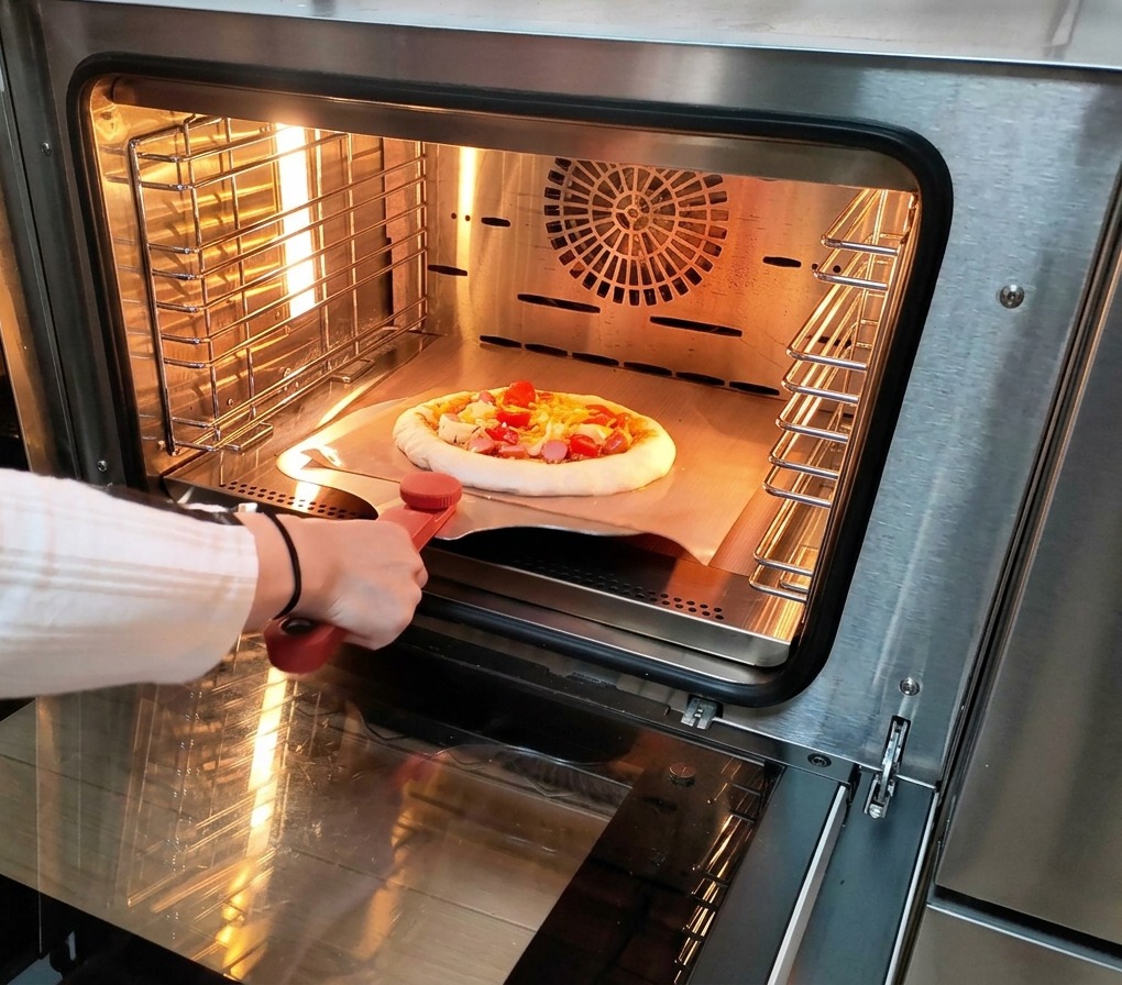 Une personne utilise une pelle à pizza métallique pour glisser une pizza crue garnie de sauce, de fromage et de viandes dans un four électrique chaud en acier inoxydable.