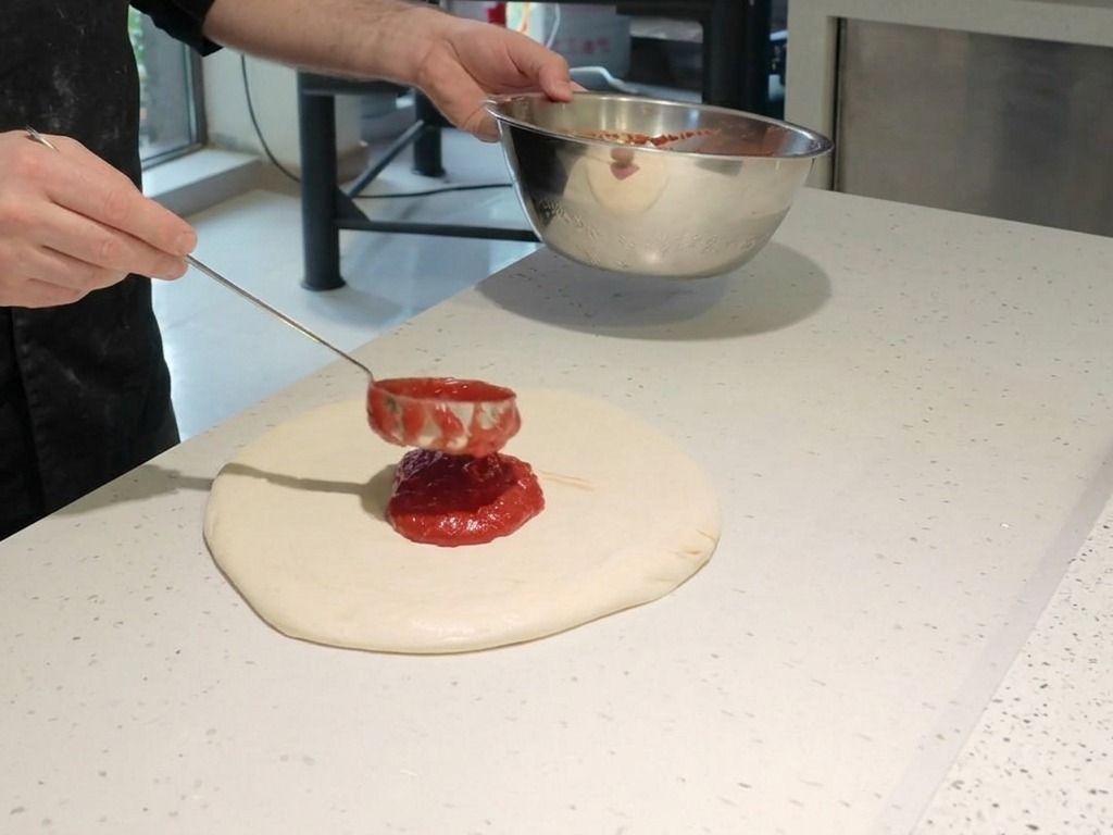 Un chef utilisant une louche métallique pour étaler une sauce tomate rouge vif en spirale sur une pâte à pizza crue.