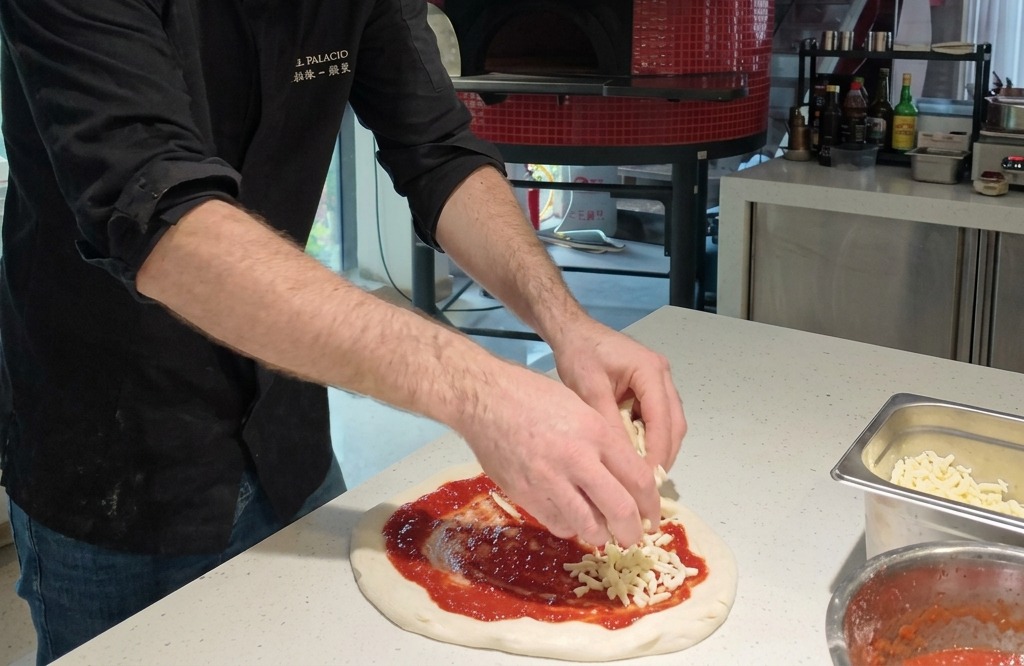 Des mains déposant des morceaux de mozzarella fraîche blanche sur une base de pizza circulaire garnie de sauce tomate.