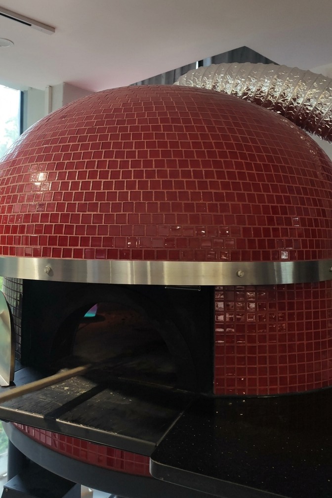 Un four à bois extérieur revêtu de mosaïque rouge alors quune pizza y est glissée à laide dune longue pelle métallique.