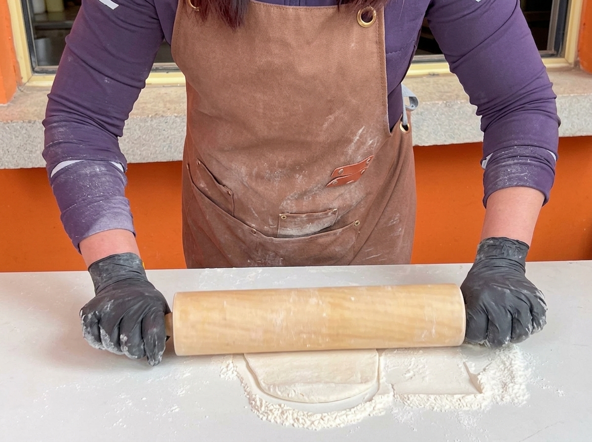 Une personne portant des gants noirs utilise un grand rouleau à pâtisserie en bois pour aplatir une base de pâte à pizza sur une table blanche farinée.