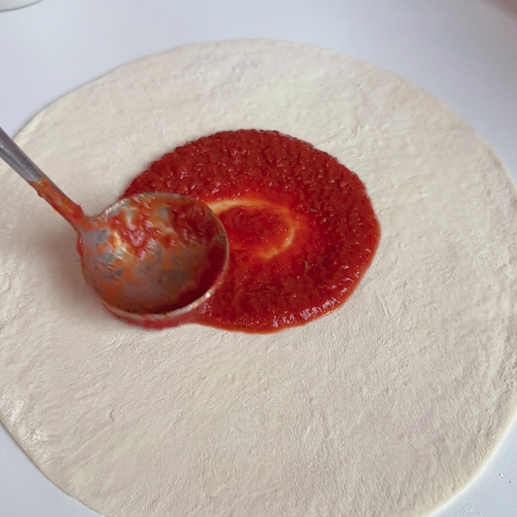 Une louche en métal répartit une sauce tomate rouge vif en un mouvement circulaire sur une base de pâte à pizza blanche et crue.
