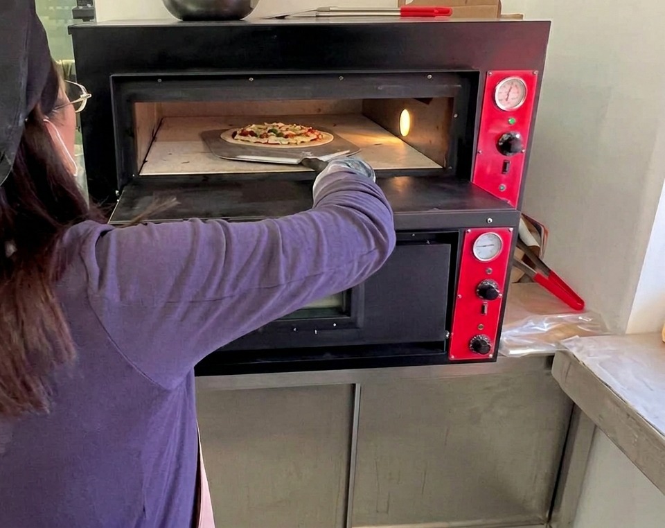 Un cuisinier utilisant une longue pelle en métal pour enfourner la pizza Margherita crue dans un four à sole électrique professionnel bien chaud.