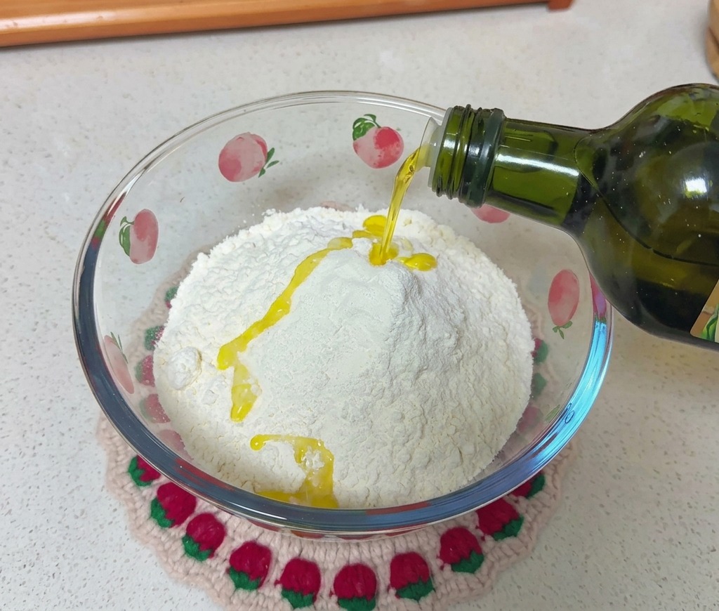 Un filet dhuile dolive versé depuis une bouteille en verre foncé sur un puits de farine blanche dans un saladier.