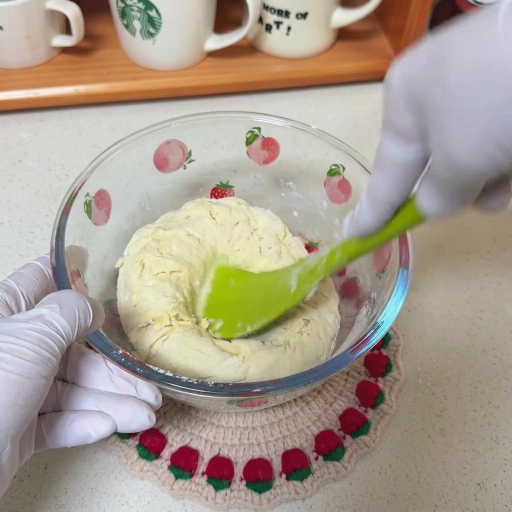 Une spatule verte mélangeant une pâte grossière de farine, deau et dhuile dans un saladier transparent.