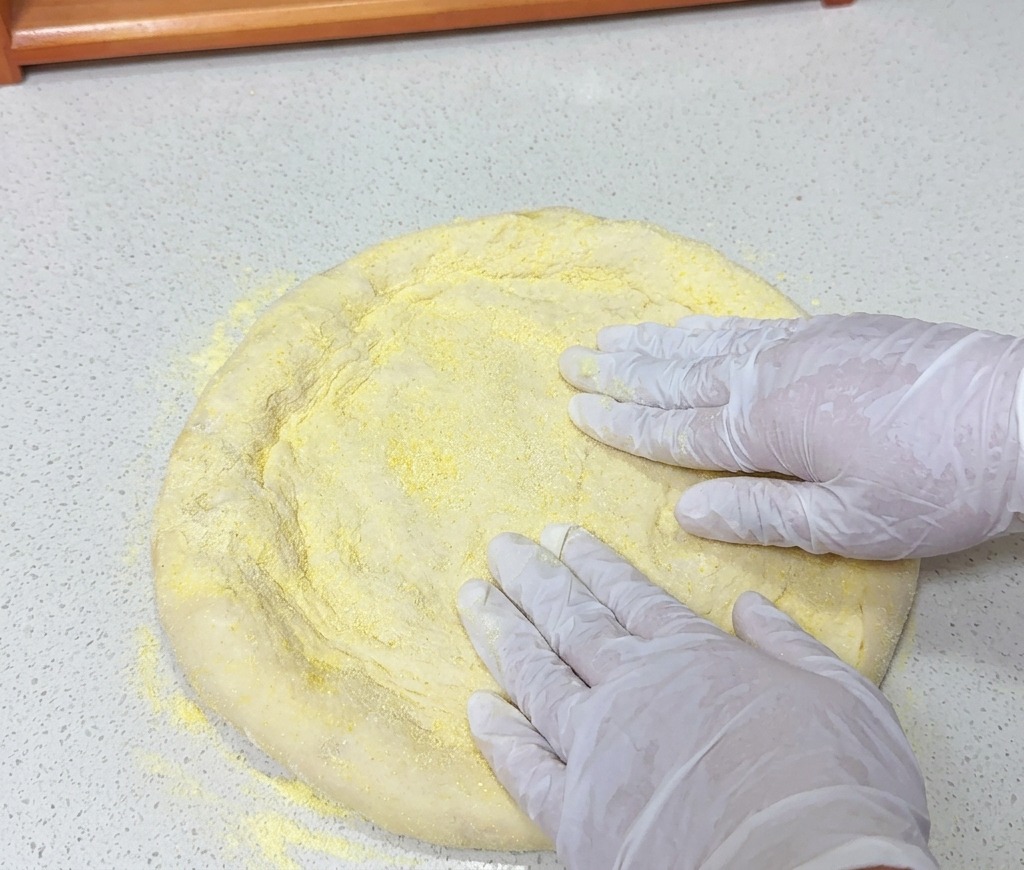 Des mains aplatissant un pâton rond enrobé de farine de maïs jaune sur un plan de travail clair.