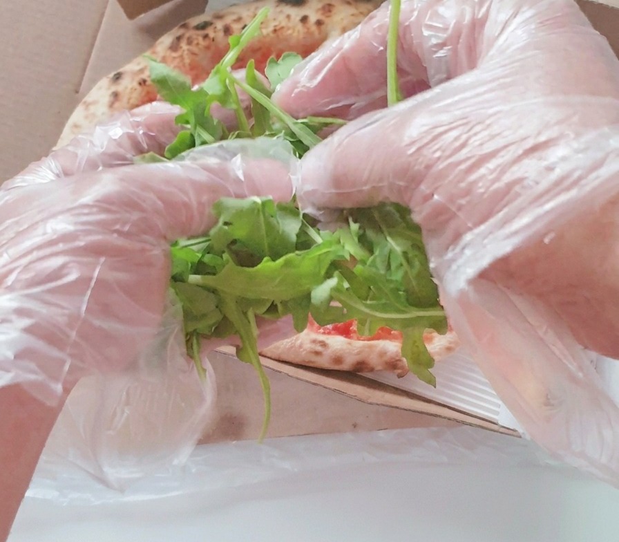 Des mains déposant une généreuse poignée de feuilles de roquette verte au centre dune pizza cuite dans sa boîte.