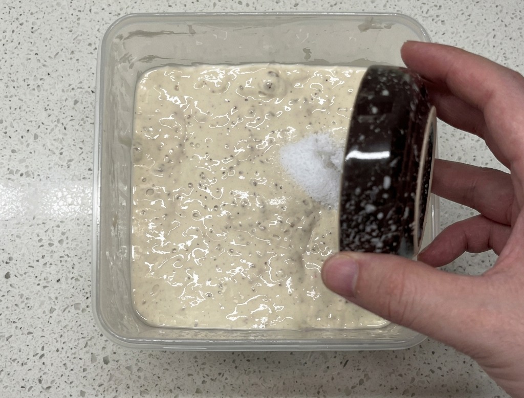 Une main versant du sel depuis un petit bol moucheté dans un récipient en plastique carré contenant un poolish pâle et plein de bulles.