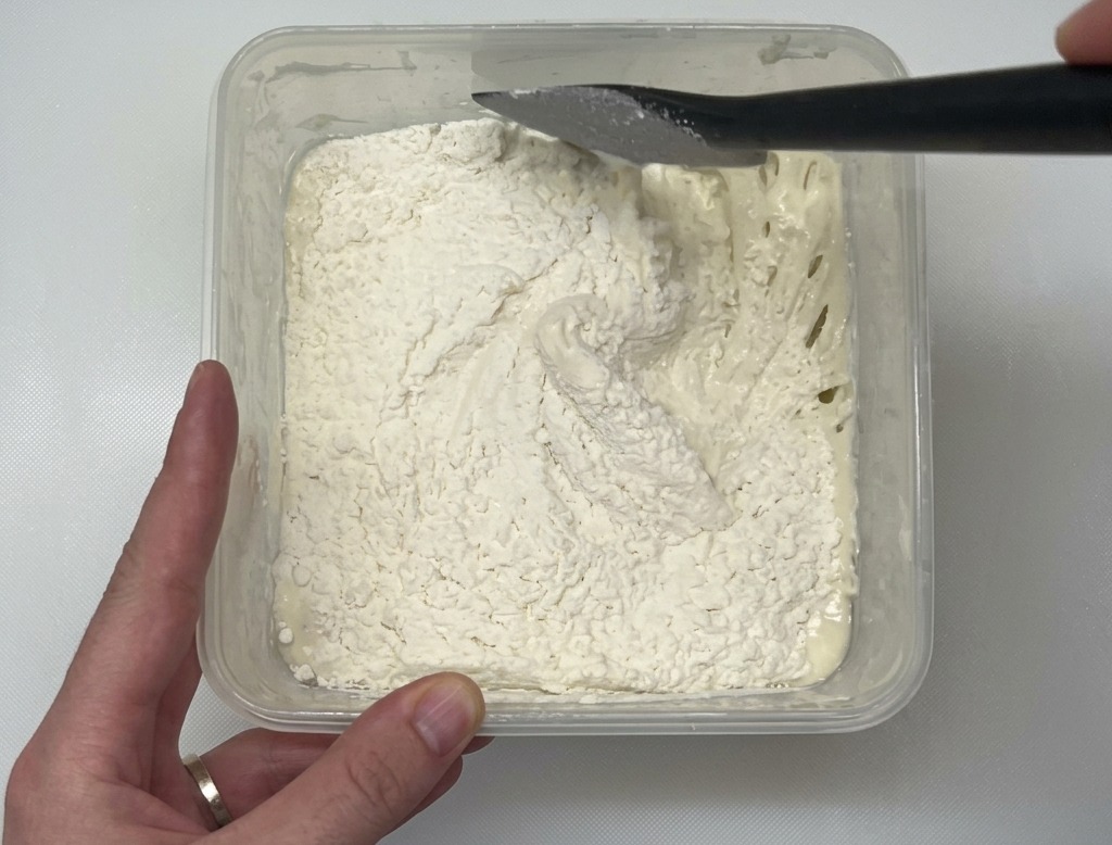 Une spatule noire mélangeant de la farine blanche dans une pâte humide à lintérieur dun récipient en plastique transparent posé sur un plan de travail blanc.